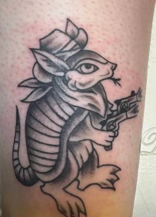 Black and gray armadillo tattoo wearing a cowboy hat and holding a revolver, depicted on skin