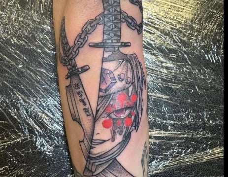 Tattoo on arm featuring a stylized face with geometric patterns, chains, and a sword design with red accents