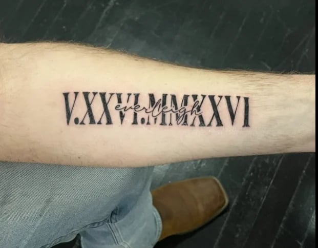 Roman numeral tattoo on forearm reading V.XXVII.MMXXVI with overlaid text