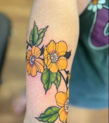 Yellow flower tattoo with green leaves on forearm, featuring blooming buds and stems in colorful ink design